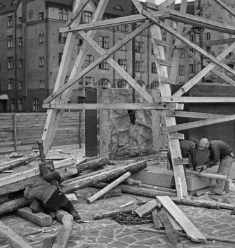 Workers erecting the Aleksis Kivi Memorial within wooden scaffolding beside stone blocks and construction materials.