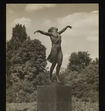 The Dancing Girl sculpture stands on a tall pedestal outdoors among trees.