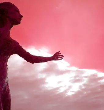 La madre del emigrante statue of a woman with an arm extended, silhouetted against a dramatic pink and white sky.