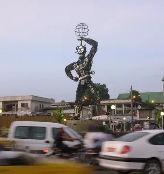 La Nouvelle Liberté towering metal sculpture in a busy street, with a spherical cage held overhead and a church spire in the background.