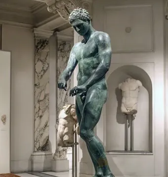 Bronze statue of the Athlete from Ephesus standing on a pedestal in a museum gallery.