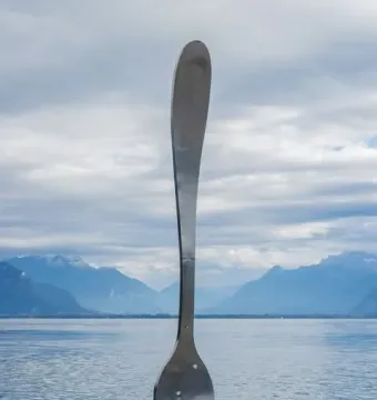 The Fork of Vevey sculpture stands upright in calm water with its reflection visible.