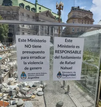 White protest signs attached to a glass barrier at the Manuel Belgrano monument, with rocks and pebbles inside the enclosure.