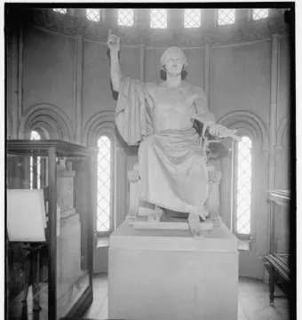 A large seated marble statue of a male figure with one arm raised and the other holding a bound object, displayed in a room with arched windows.