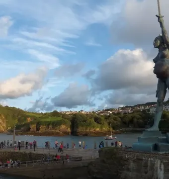 The Verity statue stands beside Ilfracombe pier with water, a grassy hillside, and a cloudy sky in the background.