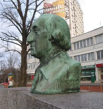 Weathered green bust of George Washington on a stone pedestal with a city street and apartment building behind it.