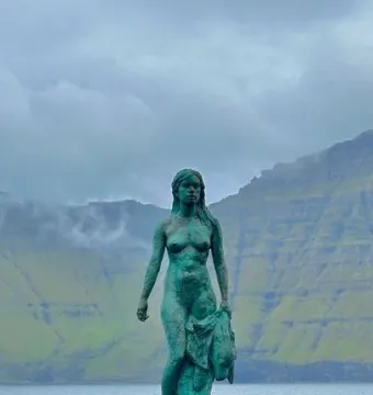 Kópakonan bronze statue standing on dark rocks by the water with misty mountains behind it.