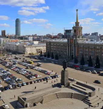Lenin Monument stands on a tall pedestal in a city square with steps, people, cars, and surrounding buildings.