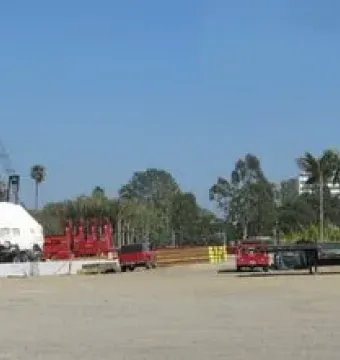 A large covered boulder for Levitated Mass sits on a flatbed trailer in a busy construction staging area with cranes, trucks, and stacked materials.