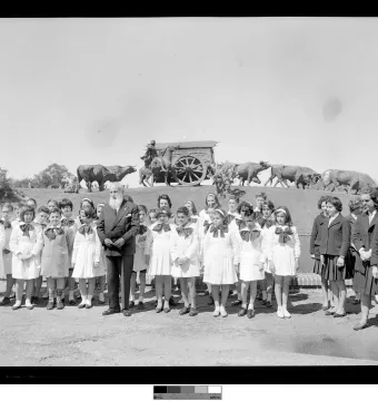 Children and an older man stand in the foreground with the Monument to the Cart on a grassy slope behind them.