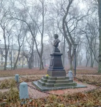Bust of Pushkin on a tall tiered pedestal in a park with bare trees and a low chain fence.