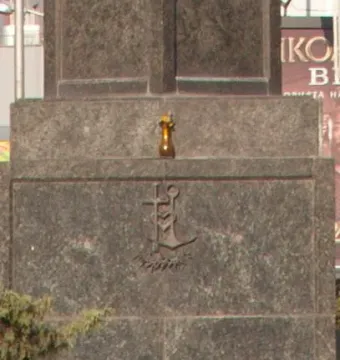 Stone base of the Statue of Sofia with a carved cross-and-anchor emblem and a small vessel on the ledge above it, partly obscured by foliage.