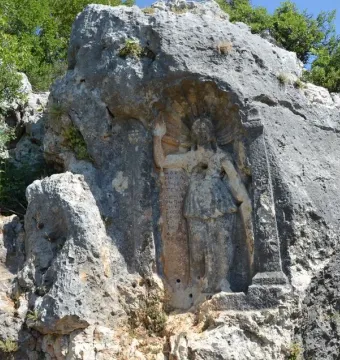 Weathered Athena relief carved into a rock face, with an inscribed panel and decorative pillar beside the figure.