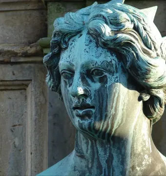 Close-up of a weathered, green-blue patinated statue head with wavy hair and a spiked headdress.