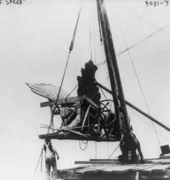 Golden Boy statue being hoisted by cables, with wooden bracing and workers below guiding the lift.