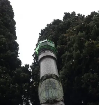 Lighthouse of Lierna, a tall stone monument with a green lantern top beside dense trees.