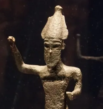 Melqart di Sciacca shown as a weathered metallic figurine with a pointed headdress and raised right arm.