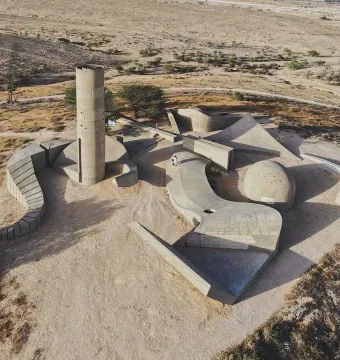 Aerial view of the Monument to the Negev Brigade, showing a concrete tower, curved walls, and geometric platforms on a dry landscape.