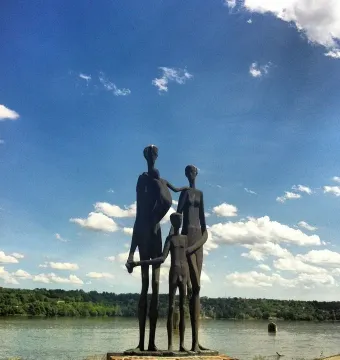 Dark bronze Monument to the Victims of Fascism sculpture of a family of five by the water under a blue sky.