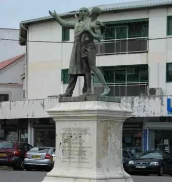 Victor Schœlcher statue with two bronze figures on a tall white pedestal at an urban intersection.