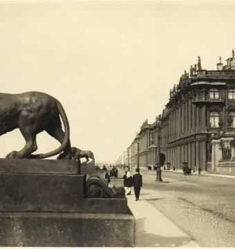 A dark lion sculpture on a tall stone pedestal at the Dvortsovaya Pier, with pedestrians and a historic building in the background.