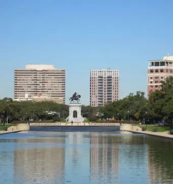 Sam Houston Statue at the end of a reflecting pool, framed by park trees and city buildings.