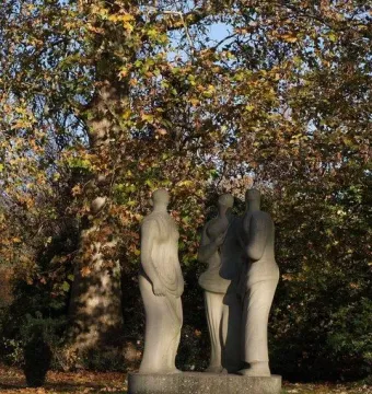 Three Standing Figures sculpture standing on a low base on a grassy lawn beside a tree.