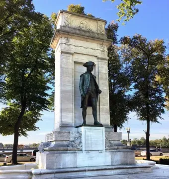 John Paul Jones Memorial with a bronze statue in a marble pylon against trees, water, and a distant bridge.