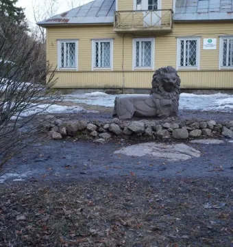 A recumbent stone lion sculpture sits on patchy ground near a yellow building, with loose rocks around it.