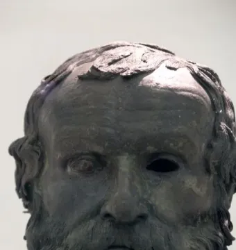 Weathered bronze head of the Porticello Philosopher with a curly beard, wavy hair, and one damaged eye socket.