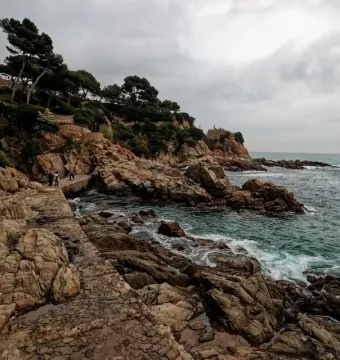 Stone stairs descend along a rocky coastline beneath a cloudy sky, with the Dona Marinera monument area in the distance.