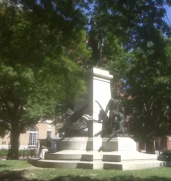 Tadeusz Kościuszko statue on a stone pedestal framed by dense green trees, with a person sitting on the steps reading.