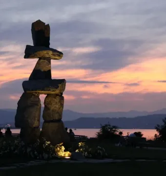 Inukshuk sculpture silhouetted at sunset beside calm water and distant mountains