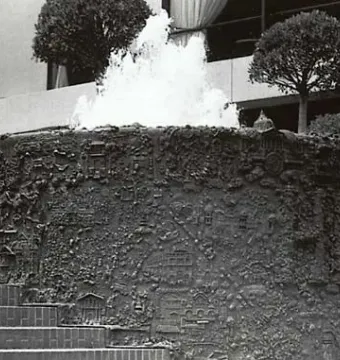 San Francisco Fountain with water cascading from its top edge in front of a modern building and two small trees.