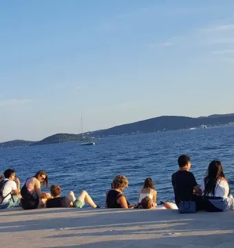 People sitting on the Sea Organ stone steps by the blue sea in Zadar, looking toward distant islands and a sailboat.
