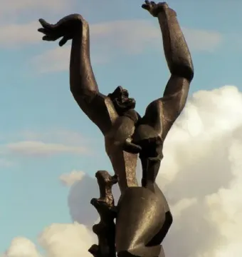 The Destroyed City bronze sculpture with raised arms and a hollow chest against a cloudy blue sky.