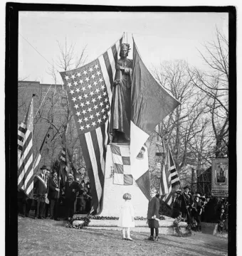 Crowned bronze statue of Dante Alighieri on a granite pedestal, with flags, wreaths, and people gathered below, including two children in the foreground.