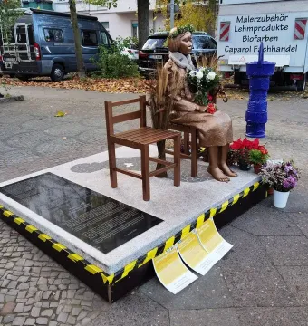 Peace Statue showing a bronze girl seated barefoot on a stool beside an empty wooden chair, holding a bouquet of flowers.