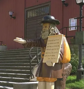 Rosenbom, a wooden statue of a bearded man in a hat and coat, holds out one hand with a scroll and bag beside him.