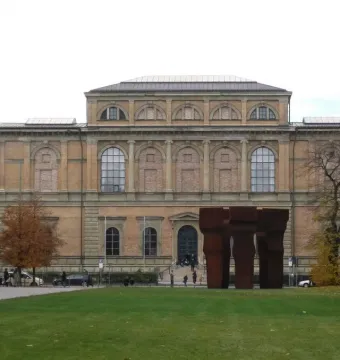 Buscando la Luz II, three tall dark reddish-brown sculptures on a green lawn in front of a classical building.