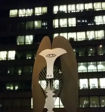 The Picasso sculpture with a stylized face and oval eyes stands illuminated at night against a building with glowing windows.