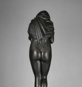 Rear view of Winter, a dark bronze female figure draped in heavy fabric and standing on a circular base.