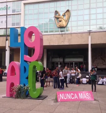 Antimonumento 49 ABC stands on a paved plaza in front of a modern building, with a small bouquet of white flowers and a pink sign reading \