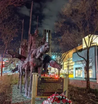 The Partisans equestrian monument at night with riders holding tall staffs, an informational plaque, and a floral wreath in front of city buildings and bare trees.