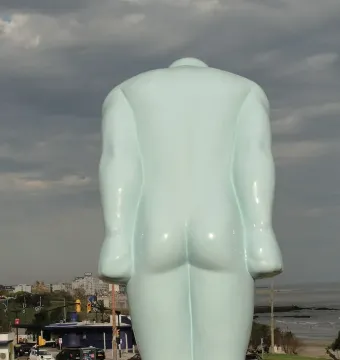 Back of the pale blue Greetingman sculpture with a cityscape, beach, and water in the background