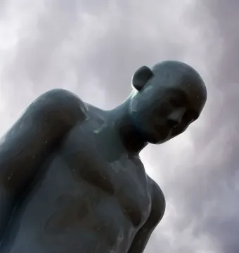 Greetingman sculpture with a bowed head against a cloudy sky.