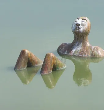 Hombre Río sculpture partly emerging from calm water, with its head tilted back and eyes closed.