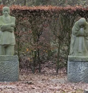 Mourning Parents stone statues by Käthe Kollwitz in a wooded setting, with one figure standing upright and the other bent forward.
