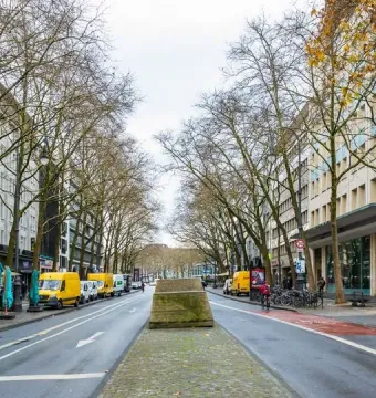 Ruhender Verkehr sculpture in the center of a wet city street in Cologne, with parked cars and bare trees along the road.