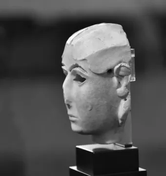 Warka Mask shown in profile on a dark stand, with hollowed eyes and an incomplete top and back.